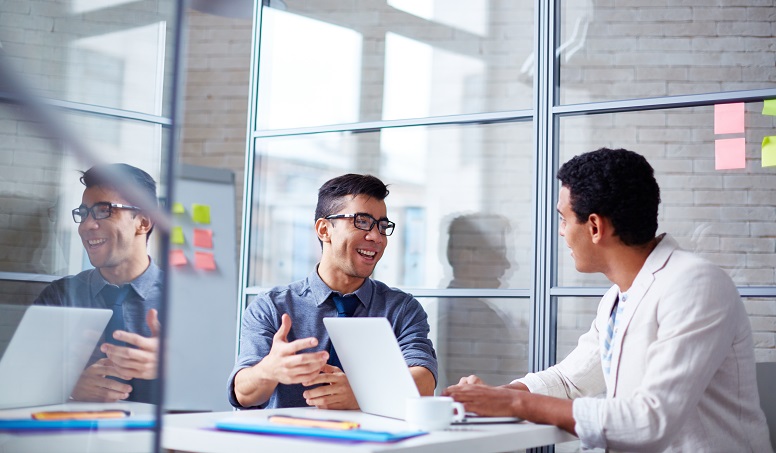Two coworkers talking while sitting at a table with one typing notes on a laptop.